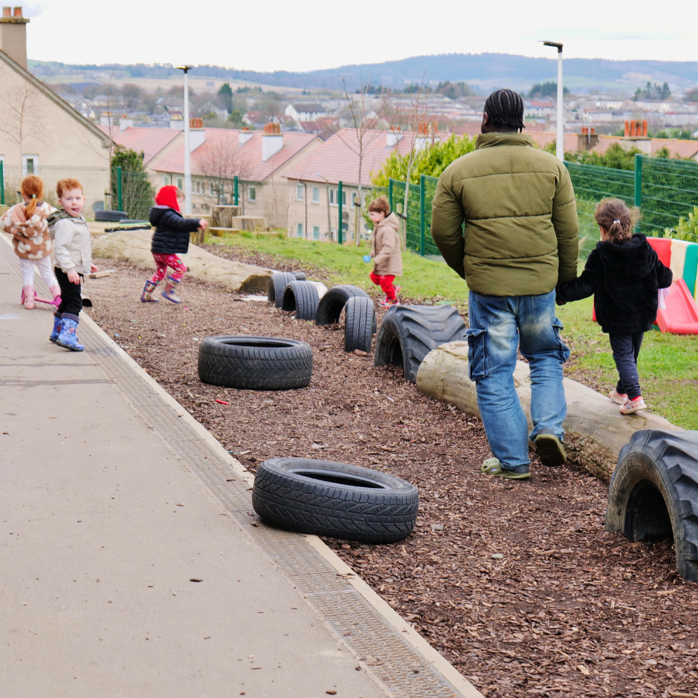 Outdoor play at a nursery, man supporting a child on a climbing frame