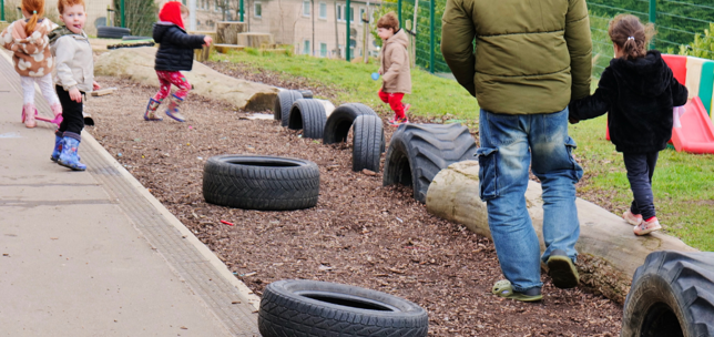 Outdoor play at a nursery, man supporting a child on a climbing frame