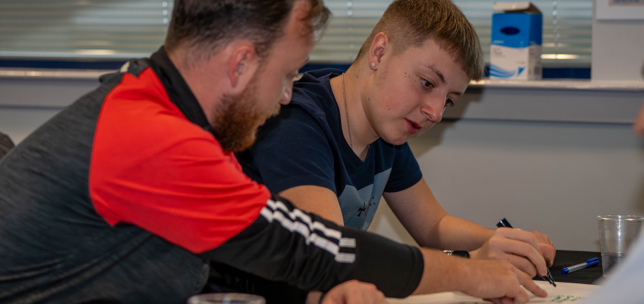 Young adult with trainer in a classroom setting, writing on paper 