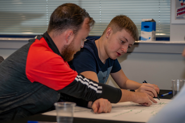 Young adult with trainer in a classroom setting, writing on paper 