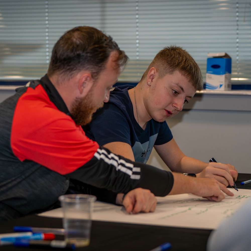 Young adult with trainer in a classroom setting, writing on paper 